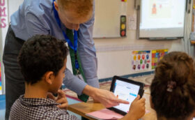 In a classroom, a boy has a tablet open in front of him. The teacher is pointing top something on it. A girl is seated next to the boy.