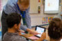 In a classroom, a boy has a tablet open in front of him. The teacher is pointing top something on it. A girl is seated next to the boy.