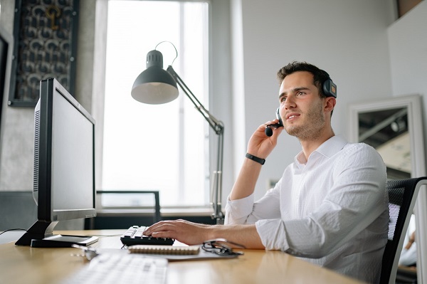 A man is sitting in front of his desktop on the table and speaking with headphones on.