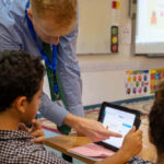 In a classroom, a boy has a tablet open in front of him. The teacher is pointing top something on it. A girl is seated next to the boy.
