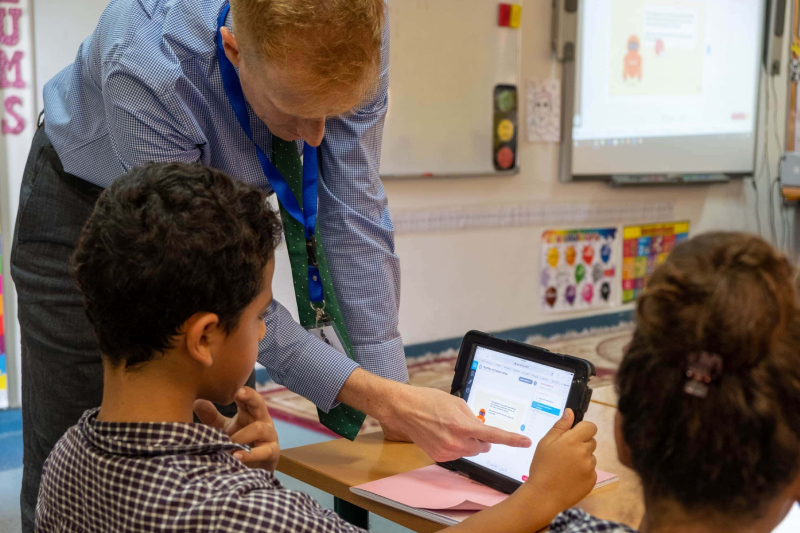 In a classroom, a boy has a tablet open in front of him. The teacher is pointing top something on it. A girl is seated next to the boy.