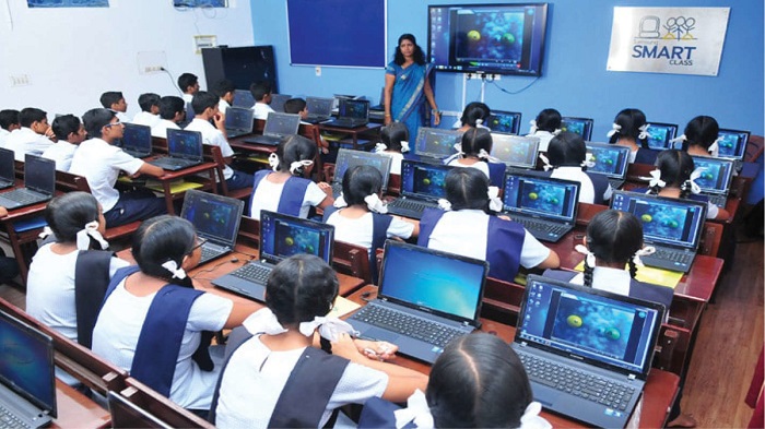 A teacher is teaching in a classroom on the projector. Students are sitting at their desks., girls on ne sde and boys on the other. A laptop is open in front of each of them. On the wall is a poster which says, Smart class.