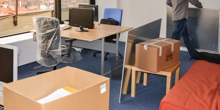 At an office environment chairs and other things are packed with plastic sheets and some workers are moving white boards from one room to another room. 